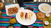 Three tacos served on a plate with salsas, toppings and tortillas