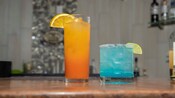 A cocktail in a tall glass with an orange garnish next to a drink with a lemon garnish and a salted rim