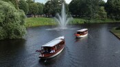 Two sightseeing boats, loaded with passengers, pass by a fountain on a river in Riga, Latvia
