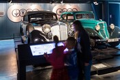 3 children reviewing a digital placard next to a display with 2 automobiles from the 1930s