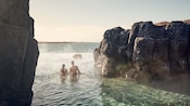Um casal entra em uma piscina com afloramentos rochosos e uma borda infinita com vista para um corpo d'água