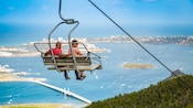 Two people riding a hillside chairlift with a lagoon in the background