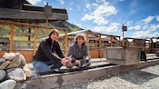 2 people smile while panning for gold in an outdoor trough with mountains behind them