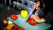 A chef slicing a bell pepper on a cutting board surrounded by various vegetables on a stainless steel countertop
