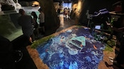 People look into a large aquarium near a painted map on a table that has text reading ‘Malta National Aquarium’