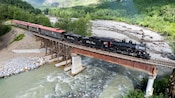 Steam train passenger cars crossing a wooden and steel railway bridge over a river, surrounded by lush hills