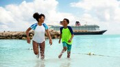 A young girl and boy smiling at each other while walking through the waters of a tropical beach with a Disney cruise ship docked nearby
