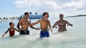 A young family of 4 playing in the waters of a tropical beach with a Disney cruise ship docked nearby