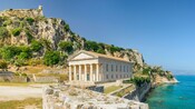 A historic waterfront site in Corfu featuring a neoclassical building with columns below a rocky hilltop with ruins