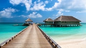 A beach boardwalk leading to thatched roof stilt houses overlooking crystal clear water