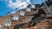 A row of gondola lift cabins traveling uphill on a wire over rocky terrain