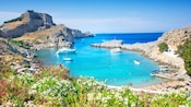 A cove in Rhodes, Greece, surrounded by rocky cliffs, with anchored boats, a sandy beach and flowers in the foreground
