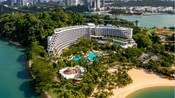 Resort con un diseño curvo cerca de la playa, con vista a un paisaje urbano con palmeras y una piscina, con las palabras “Shangri la”