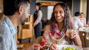 A woman laughing while drinking a beverage next to man eating a salad at Palo Trattoria on board the Disney Adventure