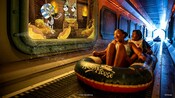 An artist rendering of 2 Guests riding the Aqua Mouse water ride past a screen showing Mickey Mouse and Minnie Mouse encountering a mummy in a tomb