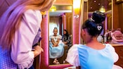 A girl looking at her reflection after receiving a princess makeover as her mother and a Cast Member look on at Bibbidi Bobbidi Boutique