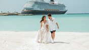 Una novia y un novio caminando por una playa con un crucero Disney a la distancia en Disney Castaway Cay