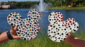 Dos personas sostienen piezas de arte en mosaico en forma de las cabezas de Mickey y Minnie frente a un lago con una fuente