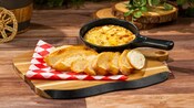 A cast iron skillet of artichoke and crab dip served on a wooden board with slices of toasted baguette