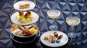A 3 tiered serving tray and a separate platter filled with small appetizers and pastries and 2 coupes of sparkling wine