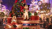 Mickey Mouse and Minnie Mouse dressed in holiday attire atop a parade float decorated with a giant Christmas tree and presents