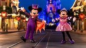 Mickey Mouse vestido como un vampiro y Minnie Mouse vestida como una princesa de hadas, parados cerca del Cinderella Castle en la noche