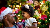 Un hombre con un sombrero de Santa Claus parado con su hijo frente a un árbol de Navidad