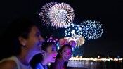 Three girls wearing Minnie Mouse ear headbands enjoying a nighttime fireworks display on a boat