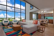 Una sala de estar de una villa grande de lujo de 3 dormitorios con un estudio y vista al Parque Temático en Bay Lake Tower at Disney's Contemporary Resort, con un sofá, 2 sillas, una mesa de restaurante, una cocina y una ventana con vistas a Magic Kingdom Park
