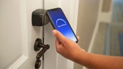 Una mano sosteniendo un teléfono inteligente que muestra una pantalla que dice ‘Door Unlocked’ (Puerta desbloqueada) en la aplicación My Disney Experience, cerca de una cerradura con entrada sin llave
