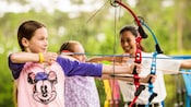 A Cast Member demonstrating proper form to 2 young girls pulling arrows back on bows