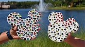 Dos personas sostienen piezas de arte en mosaico en forma de las cabezas de Mickey y Minnie frente a un lago con una fuente