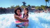 2 niñas de pie en una piscina de olas, abrazadas mientras una ola choca detrás de ellas