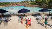 Families under beach umbrellas on lounge chairs by a pool surrounded by palm trees