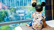 Niño pequeño con orejas de Mickey mirando por la ventana de una habitación de hotel