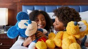 Two young kids hugging a Mickey Mouse Plush and a Pluto Plush, in their hotel room at the Disneyland Hotel.