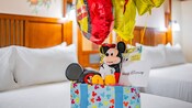 A wrapped gift, atop which is a Mickey Mouse plushie and hat, mylar balloons and a birthday card, sits on one of 2 beds in a hotel room
