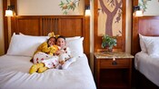 Two children hug in a hotel room bed with a painting of an orange tree and Chip and Dale on the wall