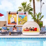 At the rooftop Pixel Pool area at Pixar Place Hotel, adults sitting in lounge chairs and 3 kids relaxing on a day bed, surrounded by vibrant artwork depicting Pixar stories