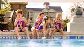 Four little girls dangle their legs into a swimming pool, splashing and laughing