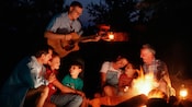 Two families gathered around a campfire while a man plays guitar