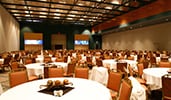 A ballroom with round tables and seating for hundreds of Guests, and a stage with twin video panels
