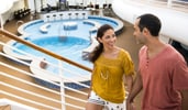 A couple holds hands while walking on a staircase with a pool in the background on the Disney Wish cruise ship