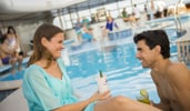 A couple sits near a pool, smiling at each other while drinking cocktails on the Disney Wish cruise ship