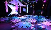 An event space with round dining tables, colorful lights, a monitor and a stage An event space with round dining tables, colorful lights, a monitor and a stage