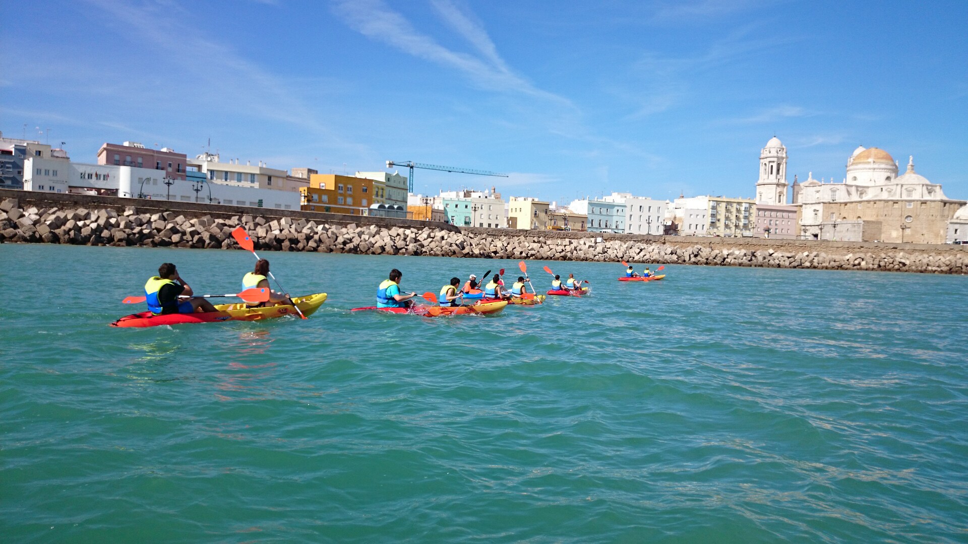 Six pairs of Guests paddle kayaks in water near a stone wall