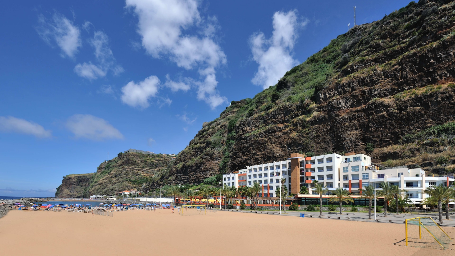Resort hotel set against a rocky cliff with sandy beach, palm trees and lounge chairs out front