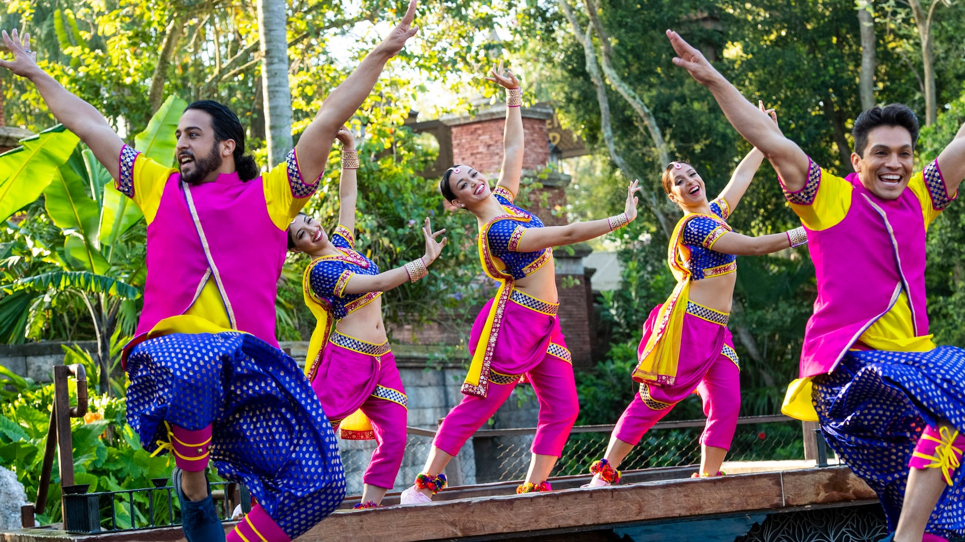 An Indian dance troupe performing in traditional dress, including 3 women dancing on stage while 2 men leap in the foreground
