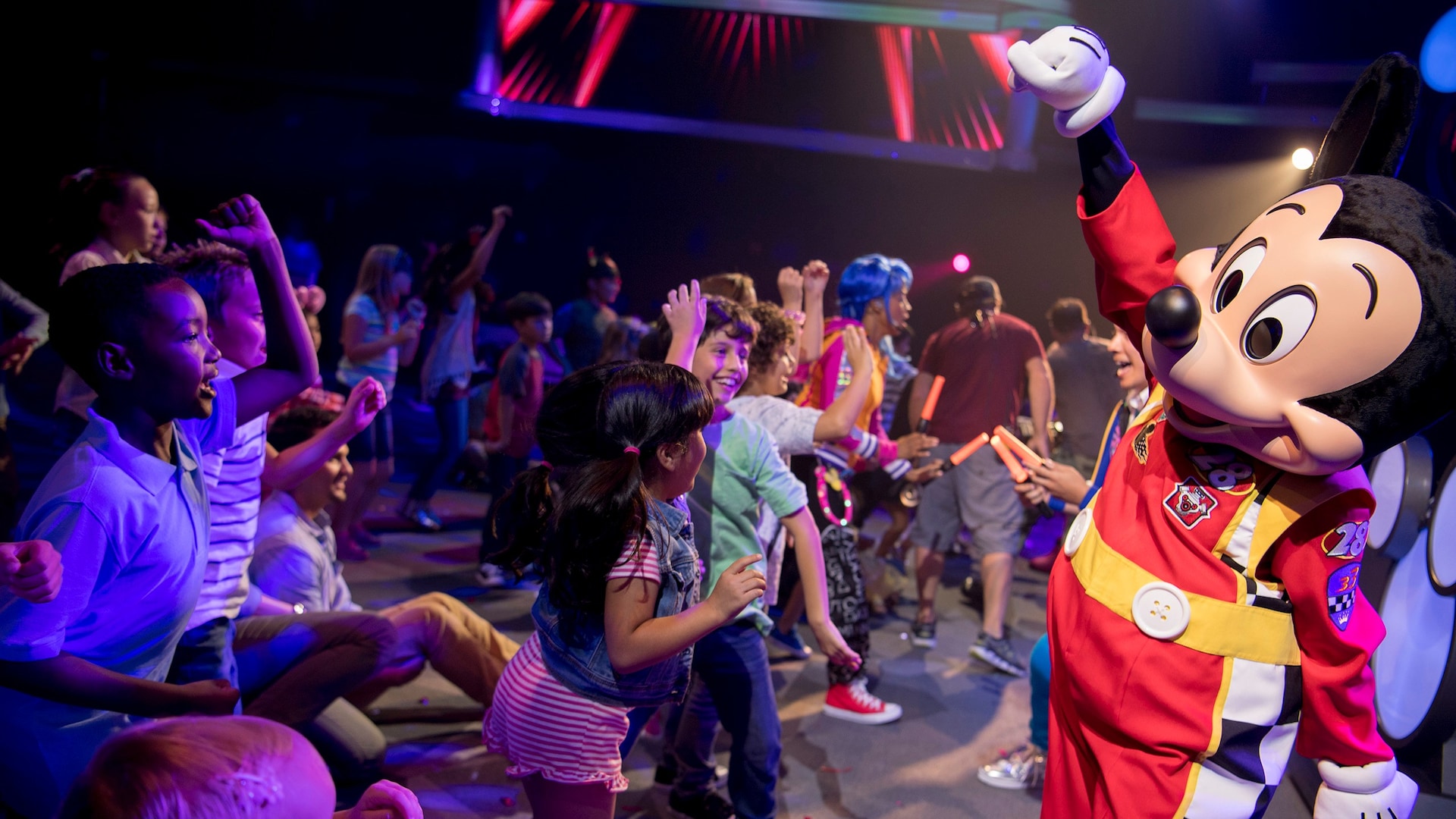 Mickey Mouse, dressed in a car racing suit, dances with children on a dance floor