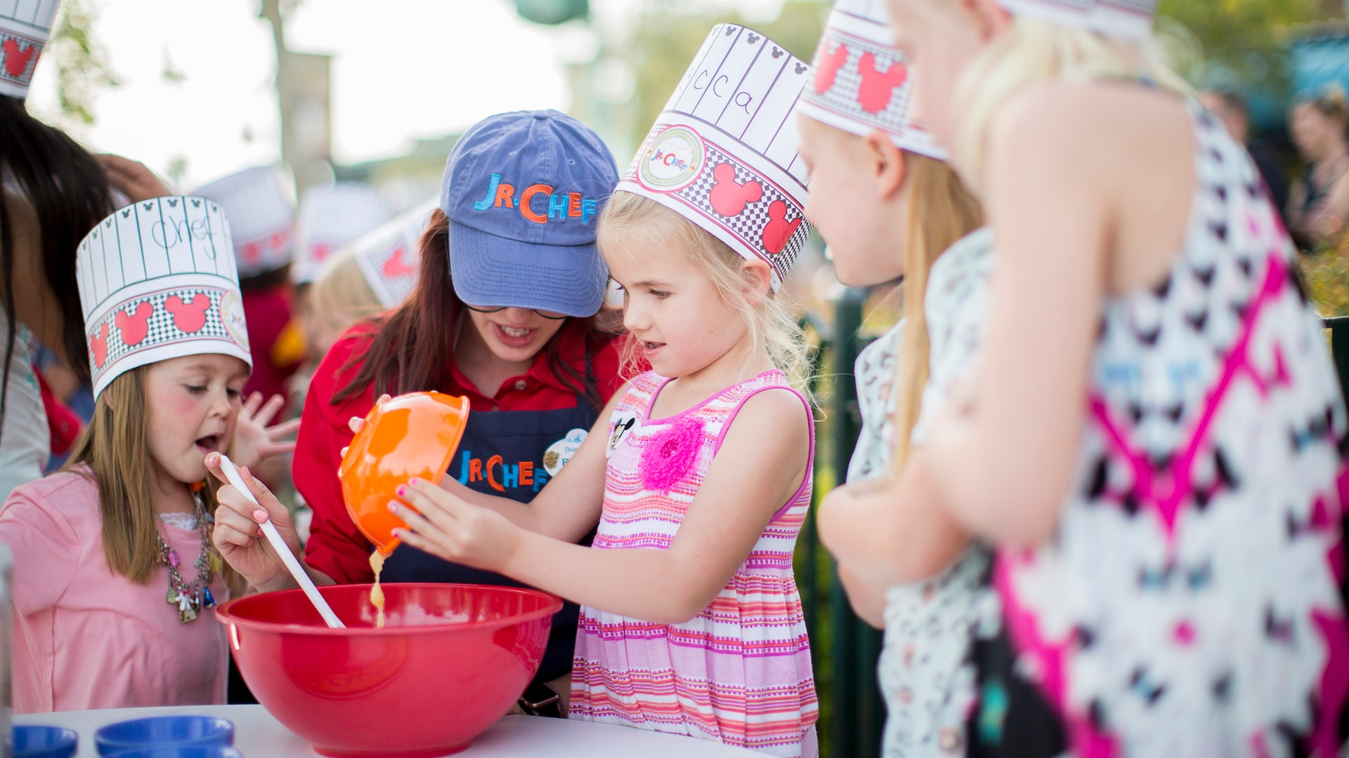 Girls wearing chef hats mix batter in a bowl with the help of a woman wearing a Junior Chef baseball cap